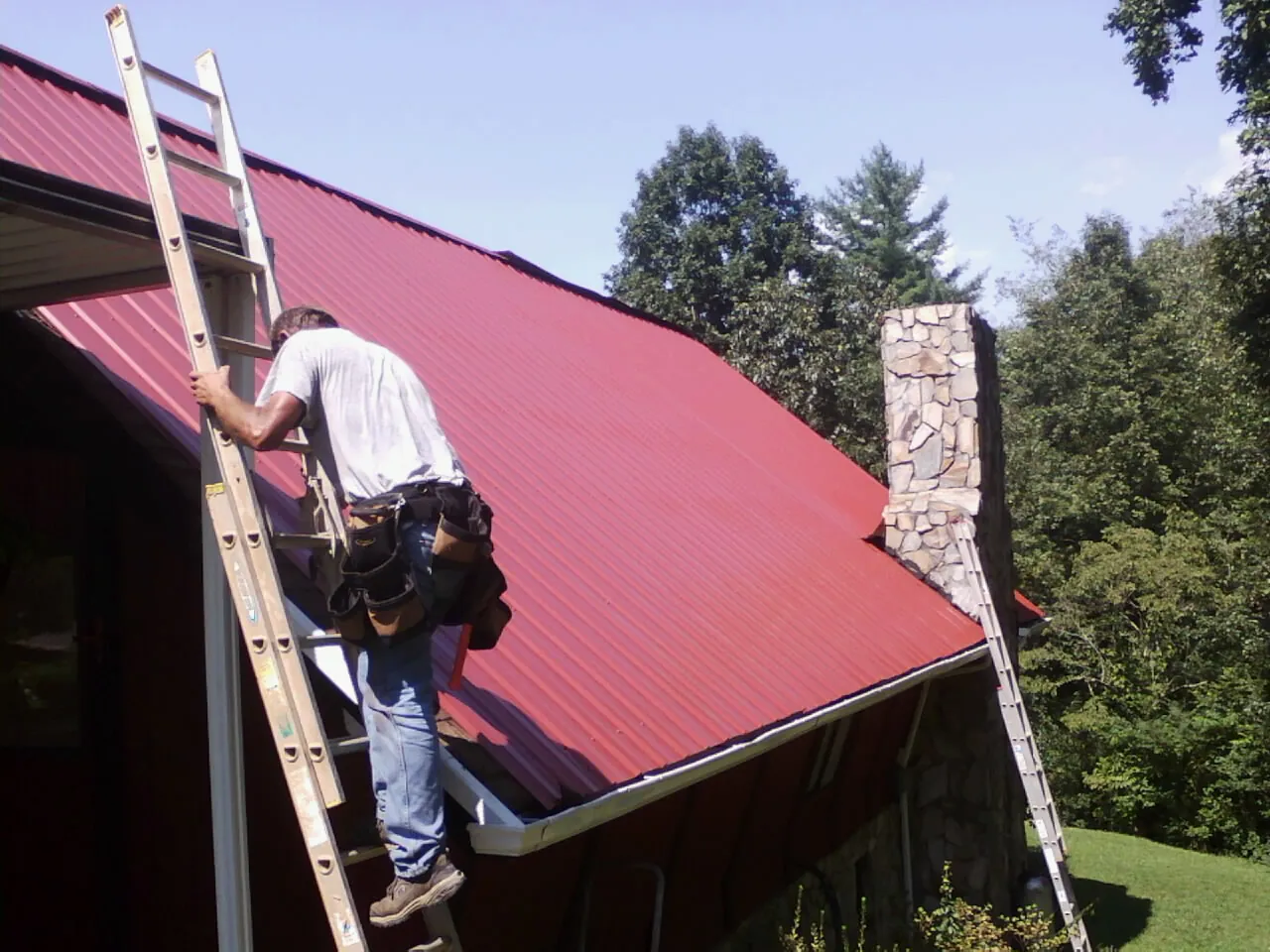 Roofer on a ladder beside a red metal roof and stone chimney — Soffit & Fascia Repair by Roofing Brothers in Sloughhouse